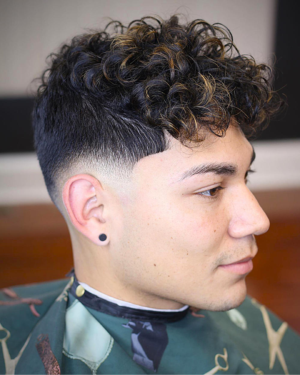 Curly top with textured waves, low skin fade, subtle highlights, clean side profile.