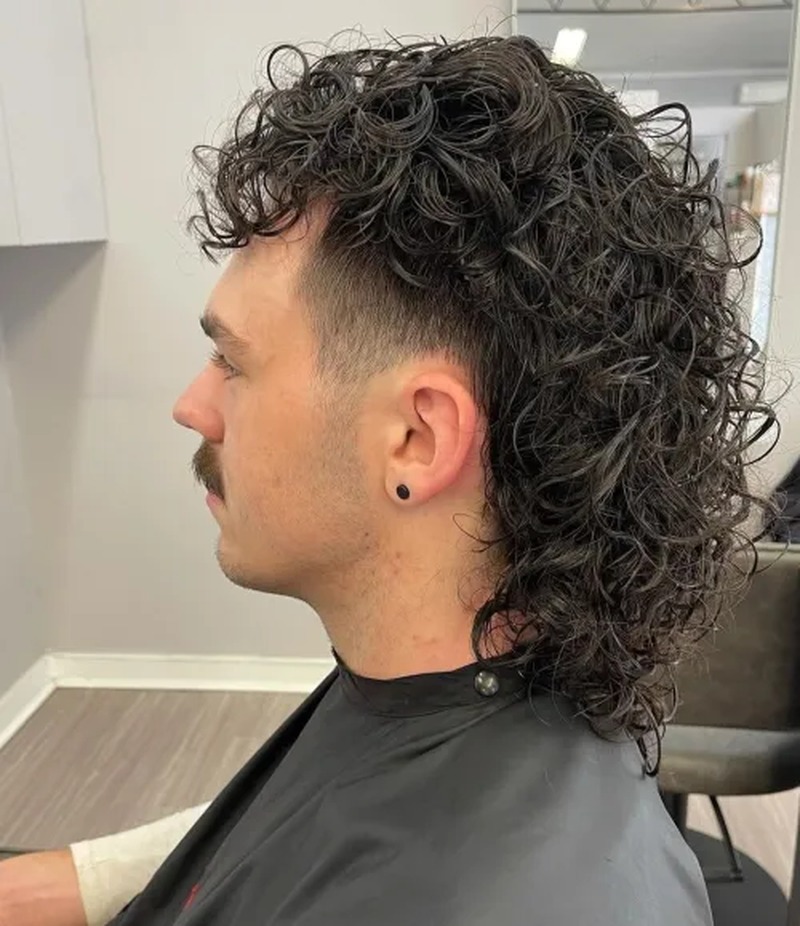 Side view of a man with curly, dark brown hair styled in a fluffy mullet variation, featuring a fade cut on the sides and voluminous curls on top and back.