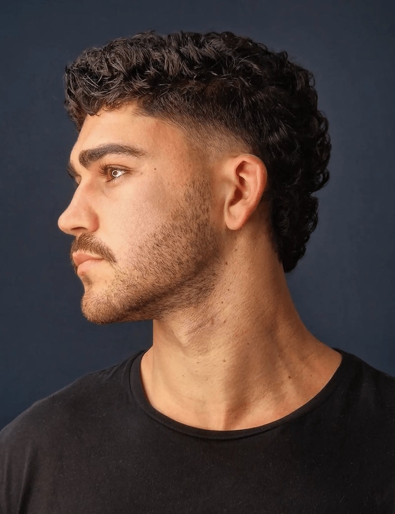 Side view of a man with curly, fluffy black hair styled in a modern mullet variation, featuring a textured cut with volume on top and shorter sides.