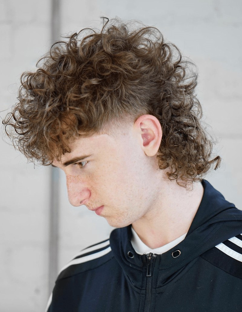 Side view of a man with curly, fluffy brown hair styled in a mullet variation featuring a fade cut.
