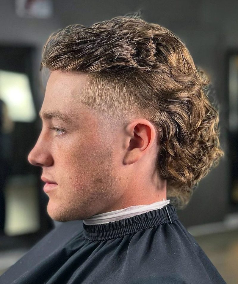 Side view of a man with curly, light brown hair featuring a fluffy mullet variation with a fade cut.