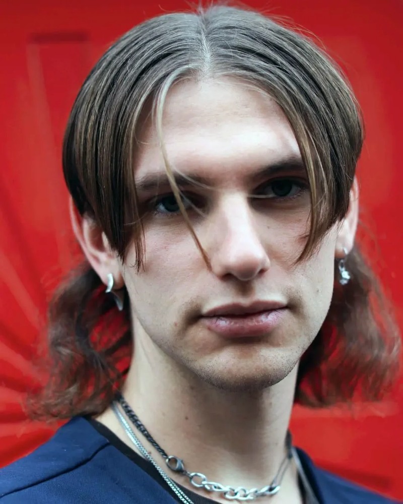 Side view of a man with medium-brown, wavy hair styled in a fluffy mullet variation, featuring a textured cut with longer, voluminous back and sides.