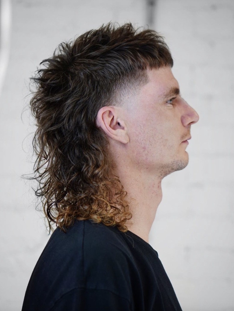 Side view of a man with wavy, fluffy mullet hairstyle featuring short sides, long curly back, and dark brown to light brown ombre color.
