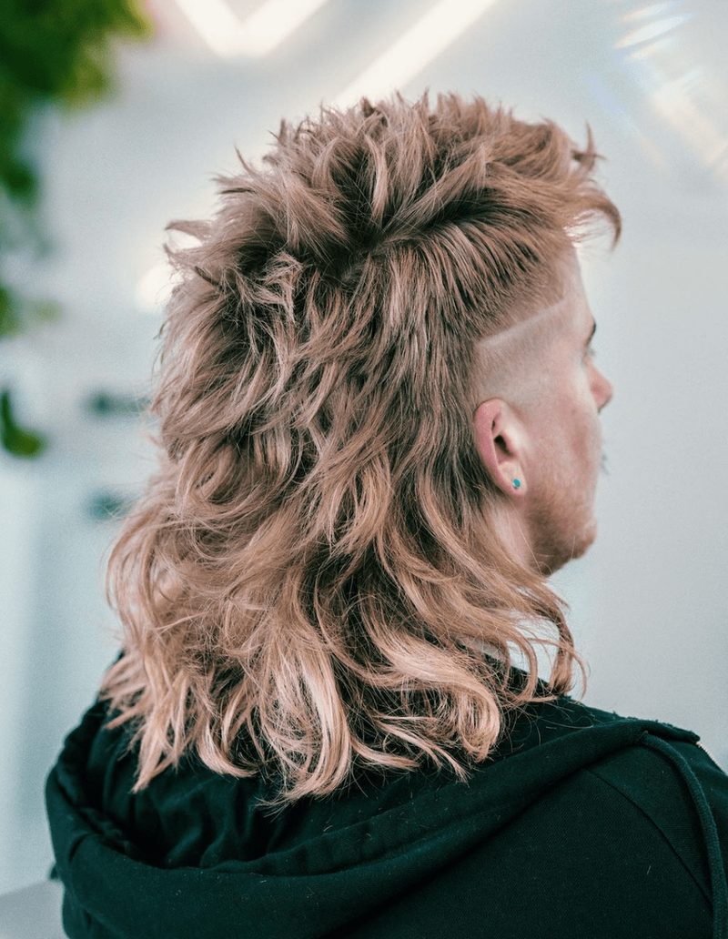 Side view of a man with wavy, light brown fluffy mullet hairstyle featuring shaved sides and textured, voluminous top and back.