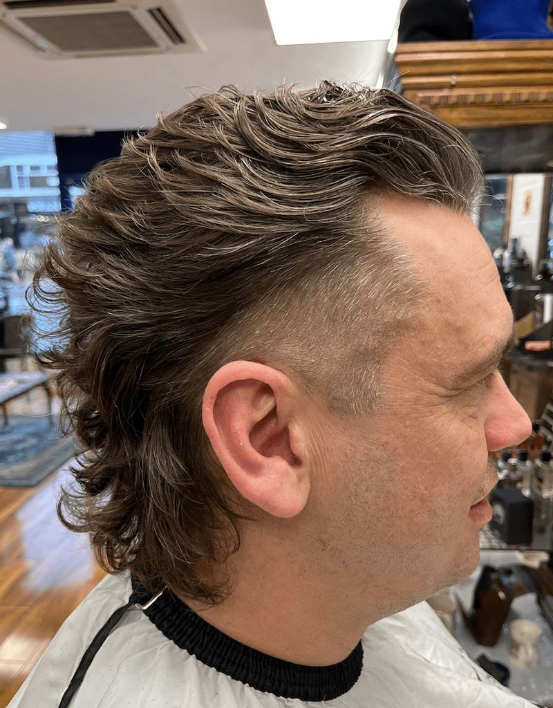 Side view of a man with wavy, medium-brown hair styled in a fluffy mullet variation, featuring a fade cut on the sides and textured, voluminous top.