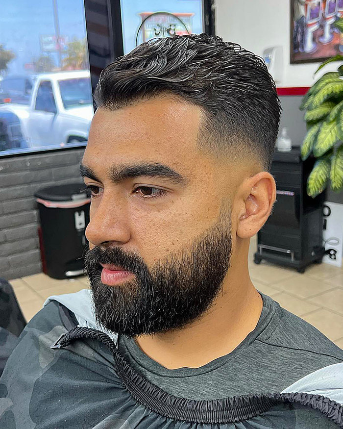 Skin drop fade with textured, swept-back top; sharp fade blends into beard, clean, modern men’s haircut.
