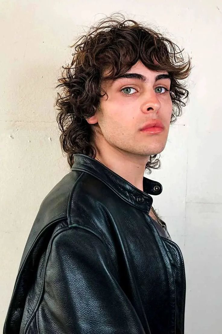 A person with a 90s grunge-inspired wolf cut, dark brown curly hair, wearing a black leather jacket, against a light wall.