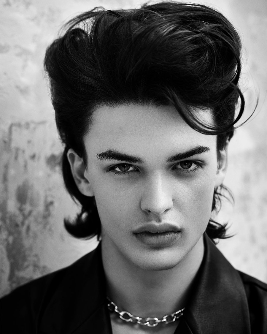 Black-and-white portrait of a person with a voluminous, dark Gothic wolf cut, wearing a leather jacket and chain necklace, against a textured wall.