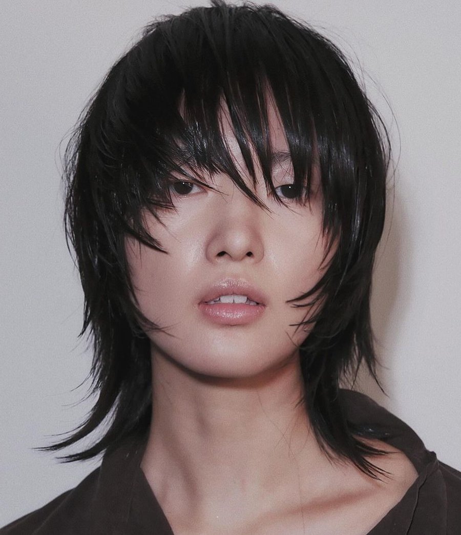 Close-up of a person with a classic K-Pop wolf cut: medium-length, layered black hair with textured, shaggy bangs, wearing a dark brown top, against a plain light background.