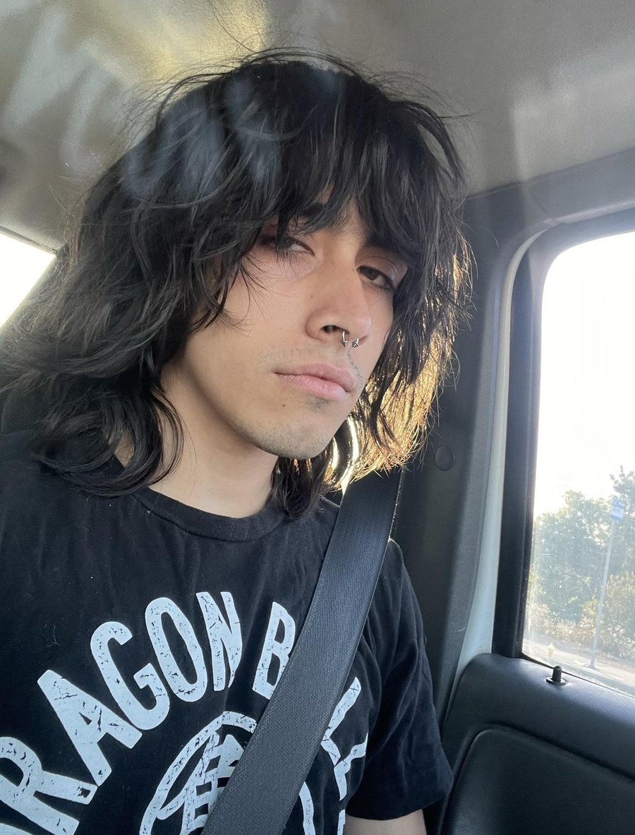 Long, wavy black wolf cut with bangs, person in car wearing black Dragon Ball shirt, natural lighting from window.